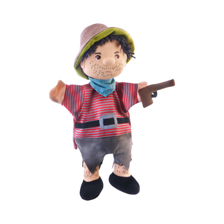 Eine Handpuppe aus Stoff, die einem Piraten ähnelt, mit braunem Hut, rot-grau gestreiftem Hemd, blauem Schal, schwarzem Gürtel, grauer Hose und schwarzen Schuhen, in der Hand eine braune Spielzeugpistole. Die Puppe hat aufgemalte Bartstoppeln.