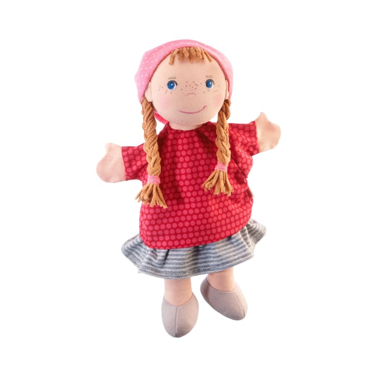 Eine Stoffpuppe mit geflochtenem Garnhaar, die einen rosa Tupfenhut, ein rotes Kleid mit Tupfen, einen grau gestreiften Rock und graue Schuhe trägt, steht vor einem weißen Hintergrund.