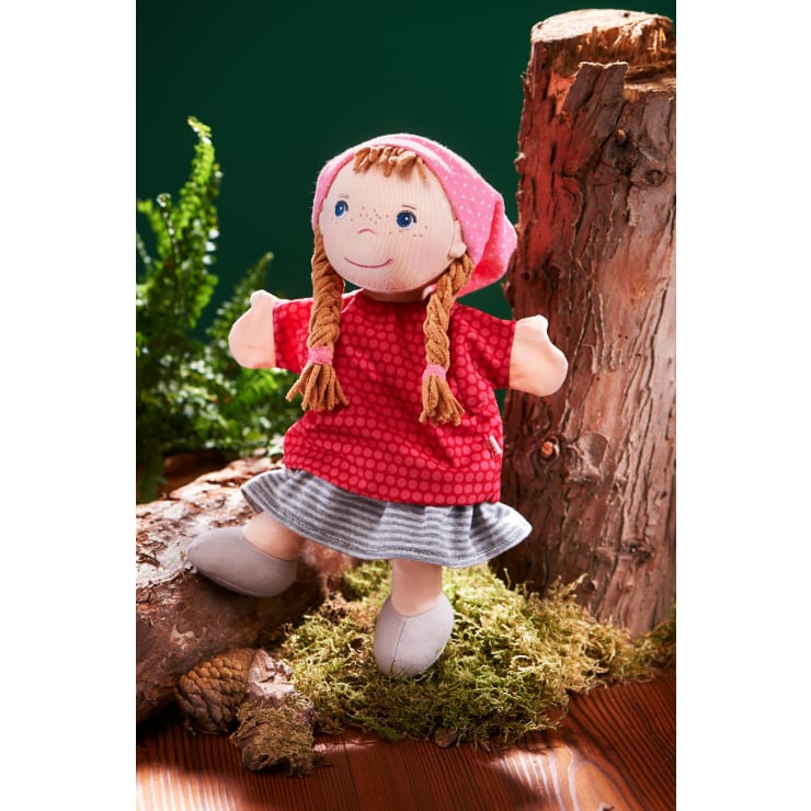 Eine Stoffpuppe mit geflochtenem blondem Haar, rosa Kopftuch, rotem Kleid und grau gestreiftem Rock steht auf moosbewachsenem Boden zwischen Baumstämmen, mit Farnen im Hintergrund.