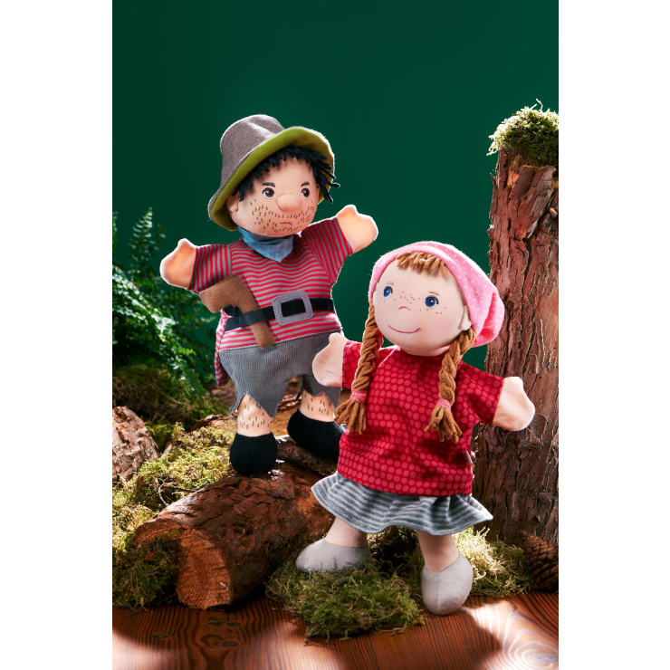 Zwei Handpuppen stehen auf einer Holzfläche mit Moos und Baumstümpfen. Eine ist als Mann mit gestreiftem Hemd, Hut und Gürtel gekleidet, die andere als Mädchen mit Zöpfen, rotem Kleid und rosa Kopftuch. Grüner Hintergrund.
