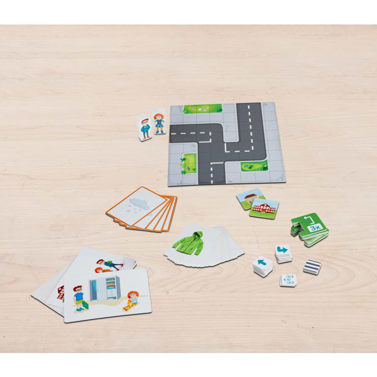 Ein Brettspiel auf einer hellen Holzoberfläche mit Straßenplättchen, Charakterplättchen, verschiedenen Karten, grünen Pappteilen, kleinen Gebäudeplättchen und drei weißen Würfeln mit Symbolen.