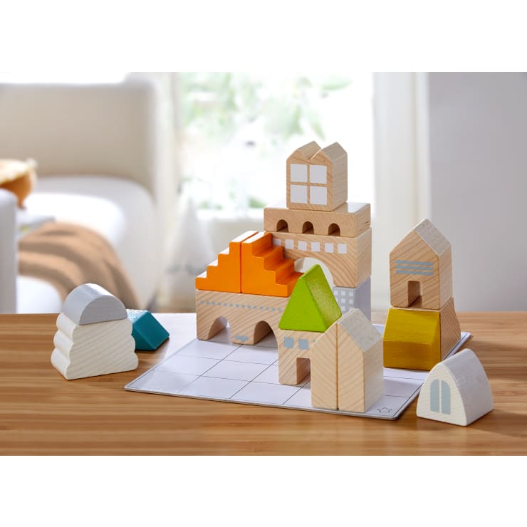 Ein Satz Holzbausteine in verschiedenen Formen und Farben ist auf einer kleinen Gittermatte auf einem Holztisch angeordnet, vor einem verschwommenen Hintergrund mit einem weißen Sofa und einem Fenster.