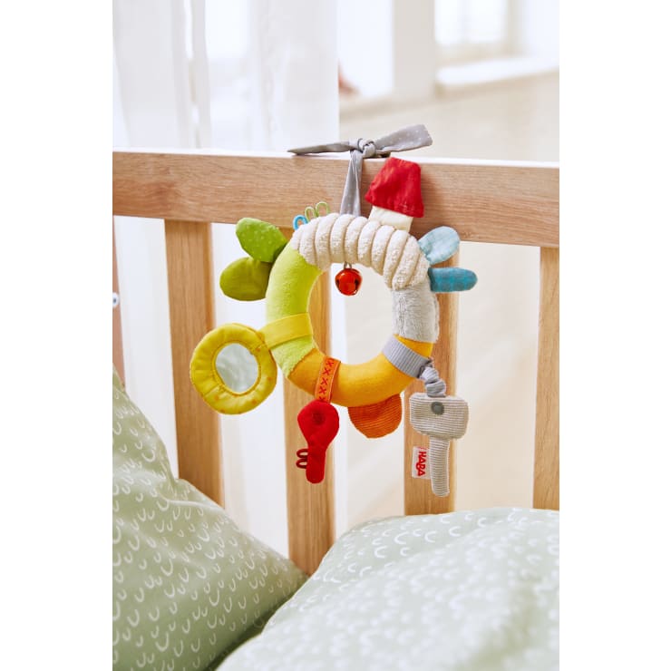 Ein bunter Babyspielzeugring aus Stoff mit verschiedenen strukturierten und geformten Aufsätzen ist mit einem grauen Band an einem hölzernen Krippengitter befestigt. Im Vordergrund ist grün gemustertes Bettzeug zu sehen.