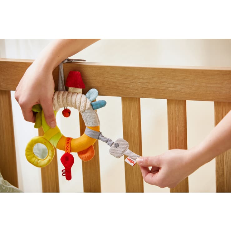 Eine Person befestigt ein buntes, kreisförmiges Babyspielzeug mit verschiedenen Stoffstrukturen und -formen mit einem Stoffband am Holzgitter eines Kinderbettes.