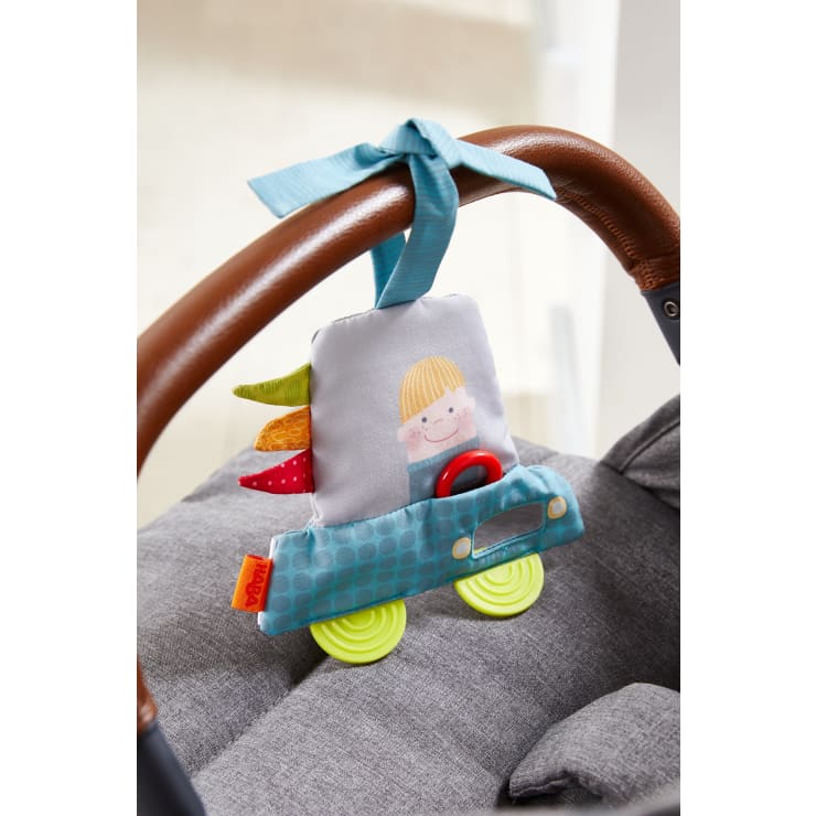 Ein weiches Stofftier in Form eines Autos mit einer lächelnden Person, bunten Anhängern und strukturierten Rädern ist mit einem blauen Band an den braunen Griff eines grauen Kinderwagens gebunden.