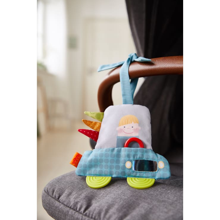 Ein weiches, blaues Plüschtier in Form eines Autos mit grünen Rädern, Stoffanhängern und einer lächelnden Kinderdarstellung auf dem Sitz ist mit einer blauen Stoffschlaufe an einem braunen Stuhl befestigt.