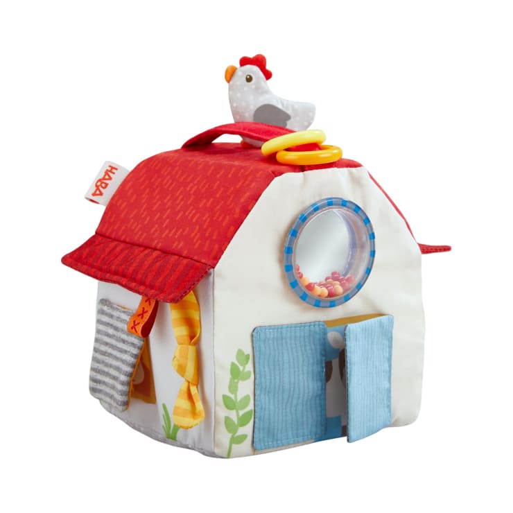 Ein Stoffspielzeughaus mit rotem Dach, blauen Türen, gelben Quasten, einem runden Fenster mit Perlen und einem Plüschhuhn obenauf. Das Haus verfügt über strukturierte und interaktive Elemente für Kinder.