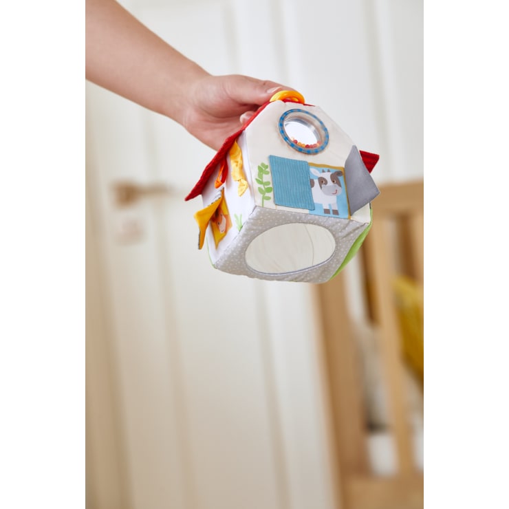 Eine Hand hält ein weiches Stoffspielzeug in Form eines Hauses mit bunten Mustern, kleinen Klappen und Tierdarstellungen, darunter eine Kuh an der Seite. Der Hintergrund ist ein Innenraum mit verschwommenen Möbeln.