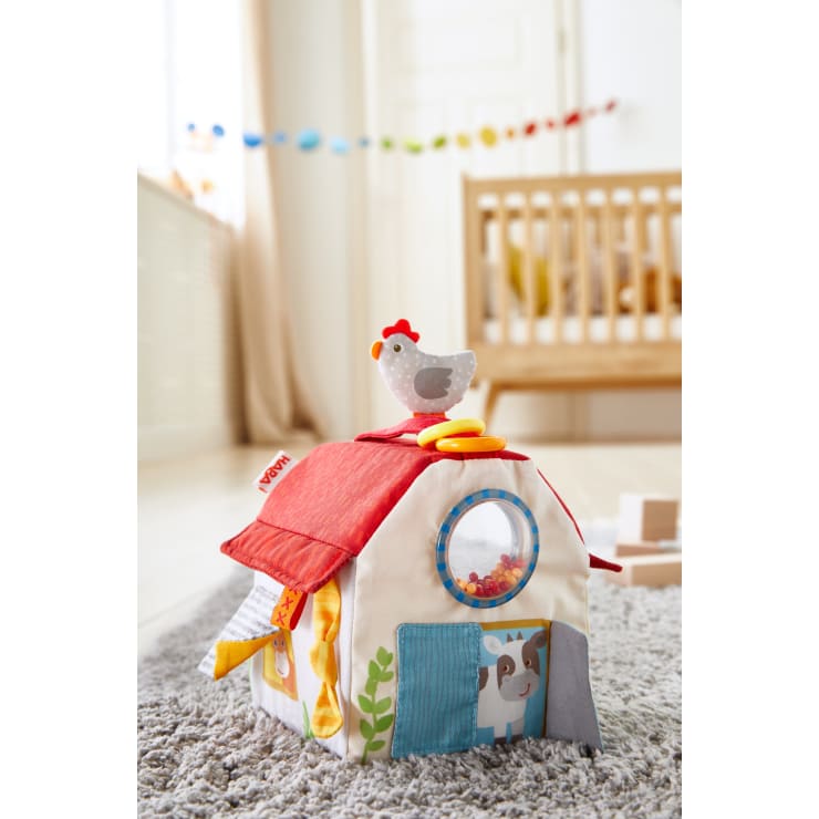 Eine Spielzeugscheune aus Plüsch mit rotem Dach, einem Plüschhuhn darauf, einem runden Fenster mit Perlen darin und bunten Türen steht auf einem grauen Teppich in einem hell erleuchteten Kinderzimmer mit einem Kinderbett im Hintergrund.