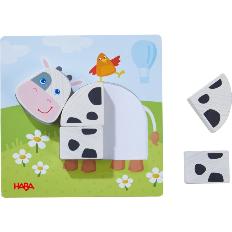 Ein Holzpuzzle mit einer Cartoon-Kuh mit zwei herausnehmbaren schwarz-weißen Teilen und einem lächelnden Gesicht. Ein roter Vogel steht auf dem Rücken der Kuh. Im Hintergrund sind Gras, Blumen und ein blauer Himmel mit einem Heißluftballon zu sehen.