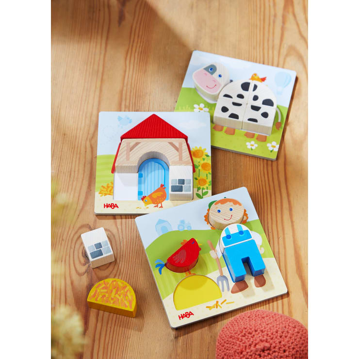 Drei farbenfrohe Holzpuzzles für Kinder sind auf einer Holzfläche abgebildet. Sie zeigen ein Haus, eine Kuh und ein Kind mit einem roten Vogel. Einige Puzzleteile sind abgetrennt und in der Nähe platziert.