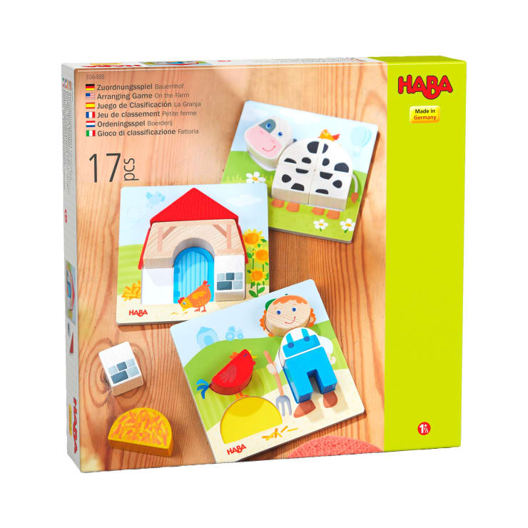 Eine Schachtel mit dem HABA-Sortierspiel "Auf dem Bauernhof" mit 17 Teilen, darunter illustrierte Holzpuzzleplatten mit Bauernhofmotiven wie einem Stall, einer Kuh, einem Bauern, einem Huhn und einem Heuhaufen.