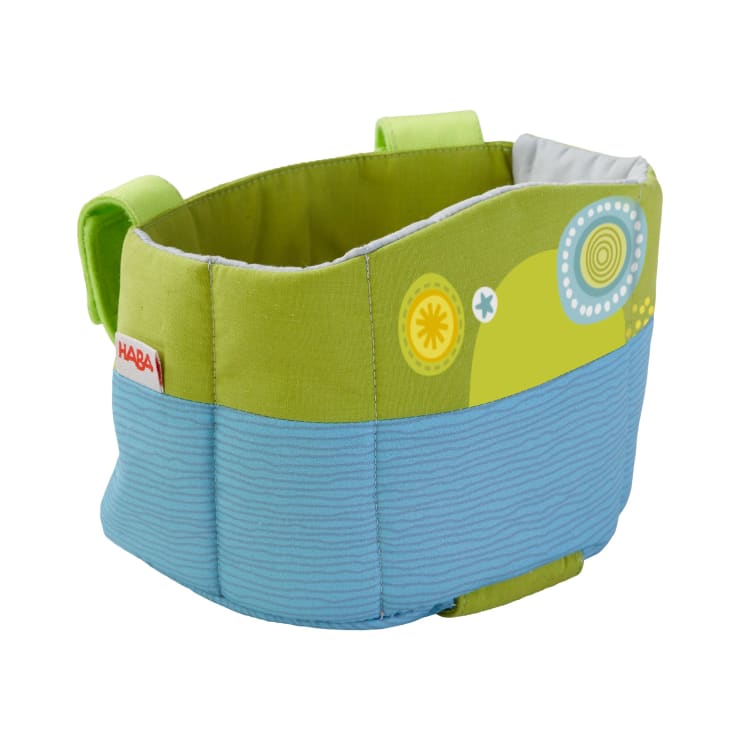Ein weicher Aufbewahrungskorb aus Stoff mit grünen Seiten, blauem Boden, dekorativen Mustern und einem roten Etikett mit der Aufschrift HABA. Der Korb hat zwei grüne Griffe und ist leer.