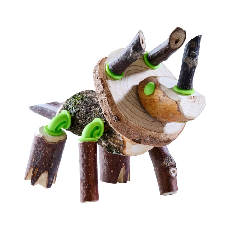 Eine kleine Tierskulptur aus Holzästen und -scheiben, die mit grünen Kunststoffgelenken verbunden sind, steht auf vier Beinen, hat einen Schwanz und zwei abstehende Teile, die an Hörner oder Ohren erinnern.