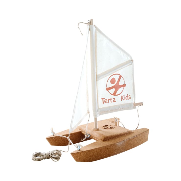 Ein kleines Modellsegelboot aus Kork mit zwei Pontons, einem weißen Segel mit dem roten Terra Kids Logo, einem Ruder am Heck und einer aufgewickelten Schnur daneben.
