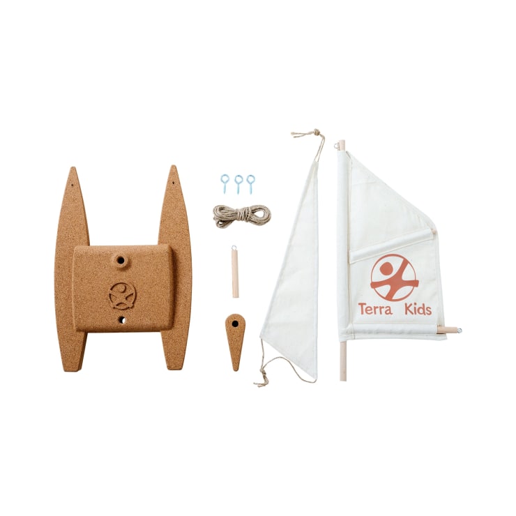 Ein zerlegter Bausatz für ein Spielzeugsegelboot enthält zwei Korkrümpfe, ein kleines Holzruder, ein weißes Segel mit dem Terra Kids Logo, drei blaue Ringe, ein Stück Seil und zwei Holzdübel, die alle auf einem weißen Hintergrund angeordnet sind.