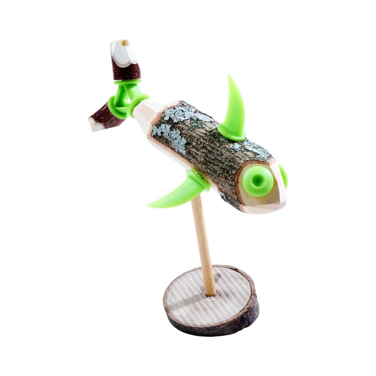 Eine handgefertigte hölzerne Fischskulptur mit grünen Flossen und Schwanz, hergestellt aus einem mit Rinde bedeckten Holzstamm und Holzteilen, montiert auf einem runden Holzsockel mit einem vertikalen Stock als Stütze, isoliert auf einem weißen Hintergrund.