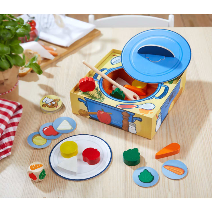 Ein Holzspielzeug-Kochset auf einem Tisch umfasst einen blauen Topf mit Deckel, einen Löffel, verschiedene hölzerne Gemüsestücke, einen Teller und runde Scheiben mit Gemüsebildern. Im Hintergrund sind eine Topfpflanze und ein rot-kariertes Tuch zu sehen.