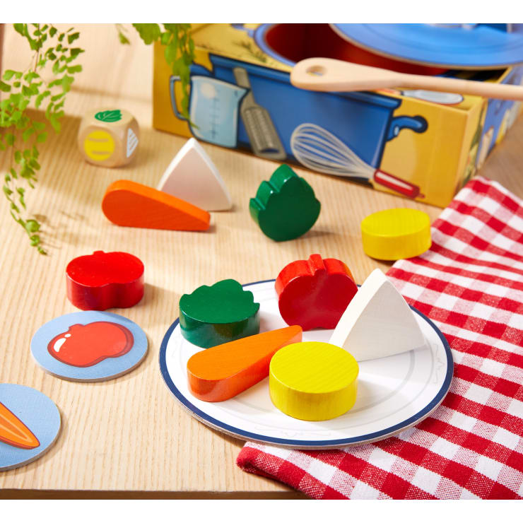 Ein Teller mit buntem Spielzeuggemüse aus Holz steht auf einem Tisch mit rot-weiß kariertem Tuch. In der Nähe befinden sich zwei illustrierte Spielsteine, ein Würfel, Kochutensilien und eine Schachtel mit Küchenmotiven. Im Hintergrund ist Grünzeug zu sehen.