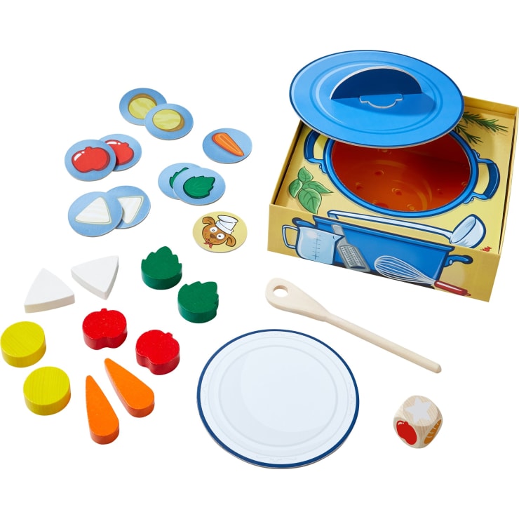 Ein Spielzeug-Kochset mit einem blauen Topf, einem Deckel, einem Holzlöffel, kreisförmigen Lebensmittelstücken aus Pappe, farbigen Gemüsestücken aus Holz, einem Teller aus Pappe und einem Würfel, ausgestellt auf einer weißen Fläche.