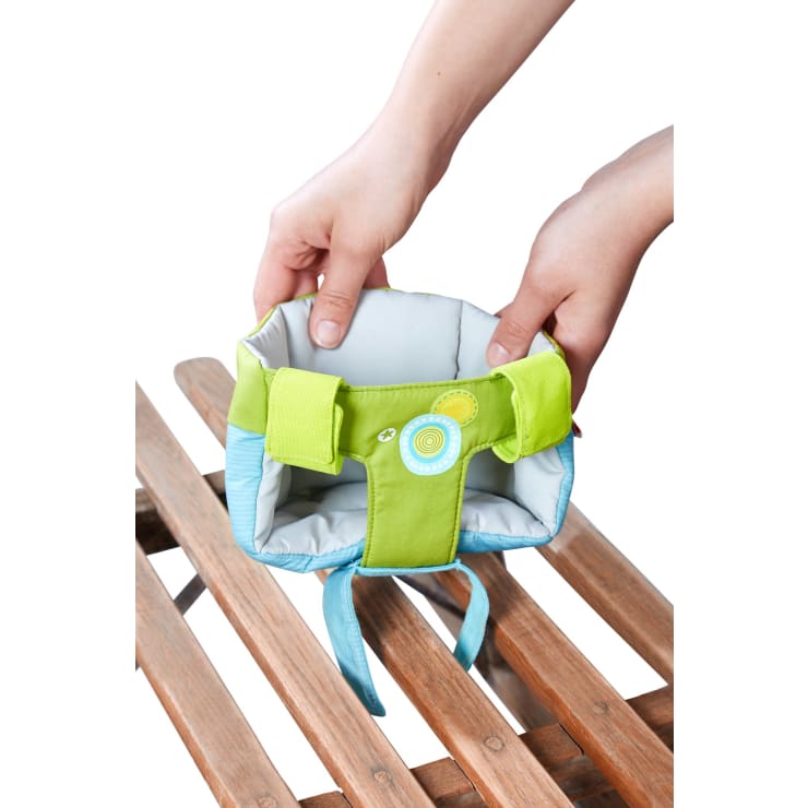 Zwei Hände halten ein kleines, gepolstertes, grün-blaues Babygeschirr mit dekorativen Kreisen über einer hölzernen Lattenfläche vor einem weißen Hintergrund.