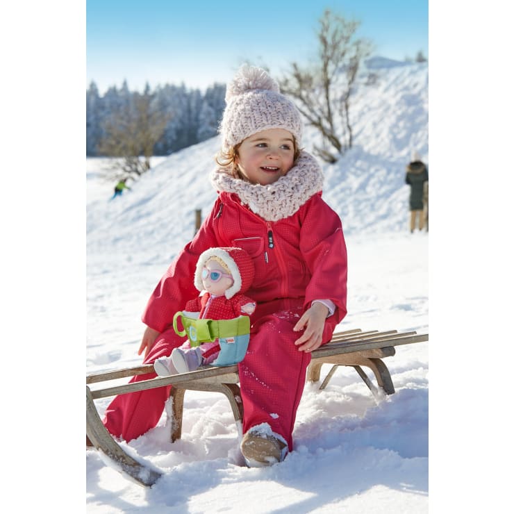 Ein kleines Kind in einem rosa Schneeanzug, einer Strickmütze und einem Schal sitzt auf einem Holzschlitten in einer verschneiten Umgebung und hält eine rot gekleidete Puppe. Im Hintergrund sind schneebedeckte Bäume und Menschen zu sehen.