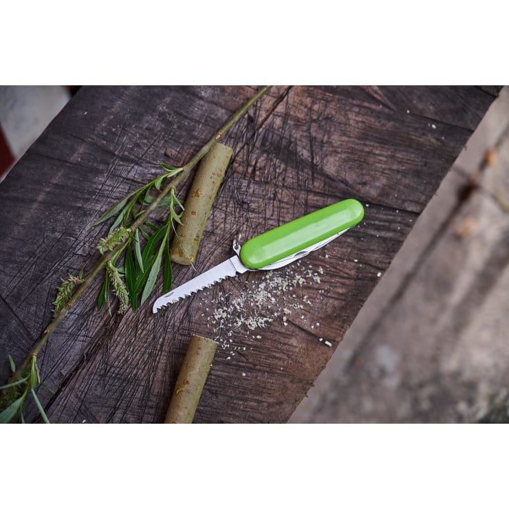 Ein grünes Taschenmesser mit gezackter Klinge liegt auf einer Holzfläche neben zwei abgeschnittenen Pflanzenstängeln, grünen Blättern und kleinen Holzspänen.