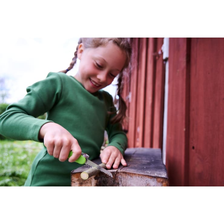Ein Kind mit geflochtenen Haaren und einem grünen langärmeligen Hemd schneidet mit einem kleinen Messer einen Stock auf einer Holzfläche im Freien, neben einem rotbraunen Gebäude.