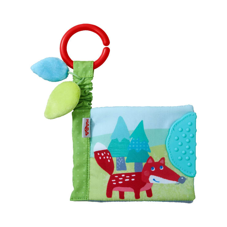 Ein weiches Babybuch aus Stoff mit einem roten Ring, zwei Plüschblättern und einer Illustration eines roten Fuchses vor grünen Bäumen. Der vordere Einband hat eine blaue, strukturierte Ecke zum Zahnen.