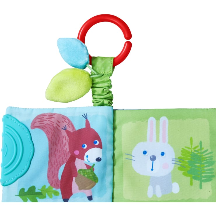 Ein weiches Babybuch aus Stoff mit einem roten Plastikring, zwei grünen und blauen Plüschblättern, einer Zeichnung eines Eichhörnchens mit einer Eichel auf der linken Seite und einem weißen Kaninchen mit Pflanzen auf der rechten Seite.