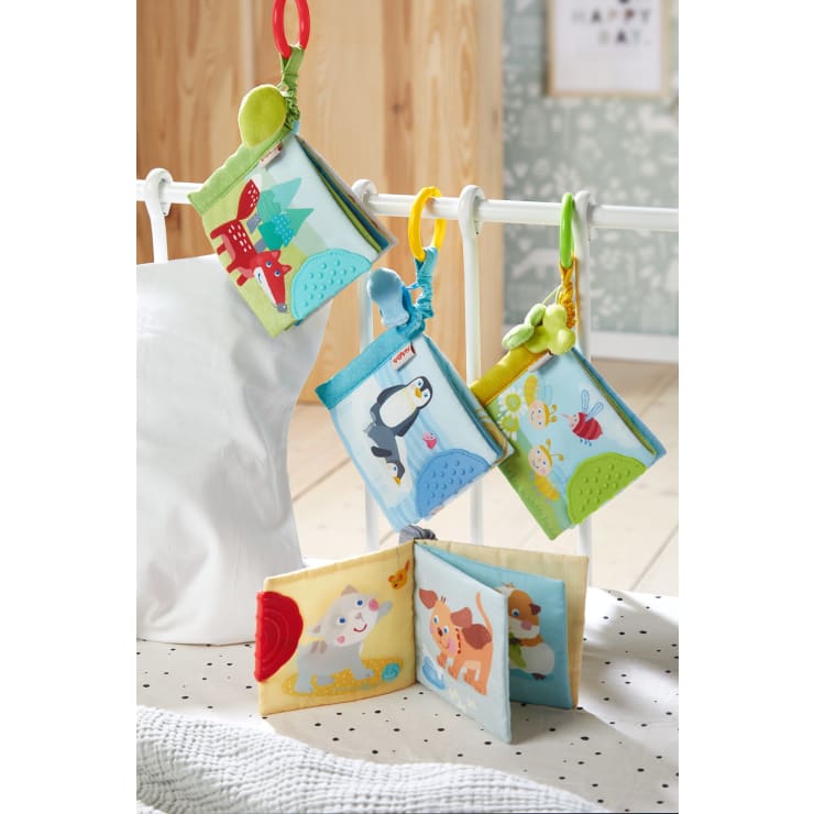 Vier Babybücher aus weichem Stoff mit bunten Tierillustrationen hängen an Klammern an einem weißen Krippengitter. Ein weiteres ähnliches Buch liegt aufgeschlagen auf einer gepunkteten weißen Fläche im Vordergrund.