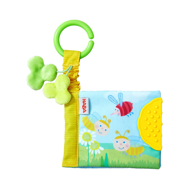 Ein weiches Babybuch mit einem farbenfrohen Einband, der lächelnde Bienen, einen rosa Schmetterling und Blumen zeigt. Es hat einen grünen Kunststoffring mit einem Plüschklee und einer gelben, strukturierten Ecke zum Zahnen.