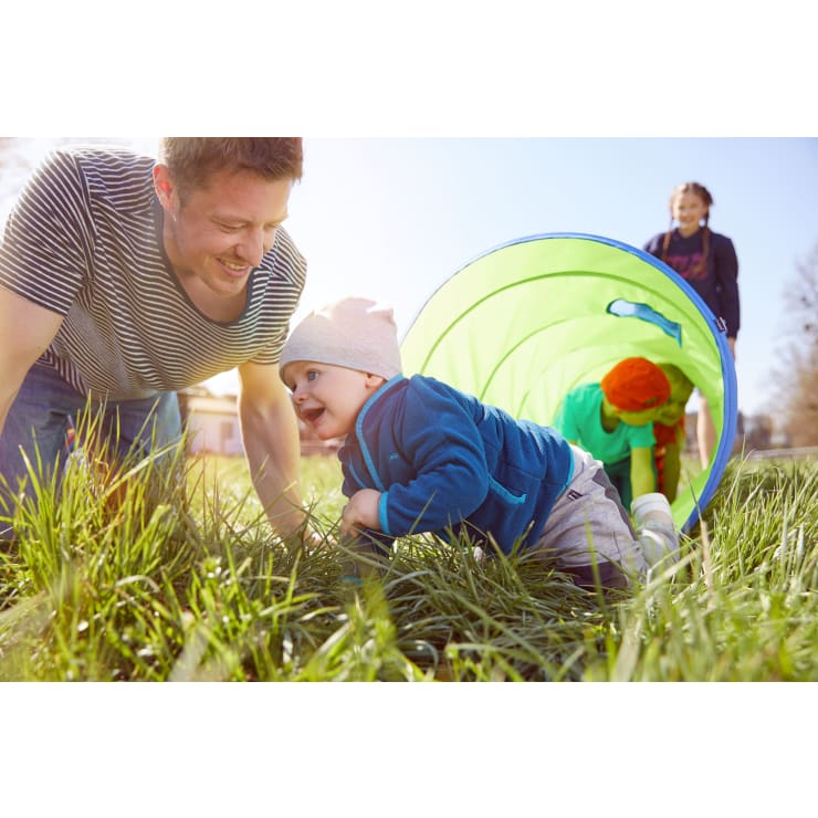 Ein Mann und ein Baby krabbeln im Gras neben einem grünen Spieltunnel. Ein anderes Kind krabbelt in den Tunnel hinein, während ein Erwachsener im Hintergrund steht. Die Szene spielt sich an einem sonnigen Tag im Freien ab.