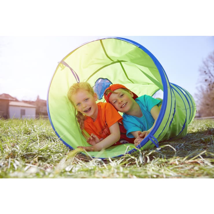 Zwei Kinder lächeln, während sie in einem grünen Spieltunnel auf einer Wiese im Freien liegen. Beide tragen bunte Hemden, und die Sonne scheint, so dass eine helle, fröhliche Szene entsteht.