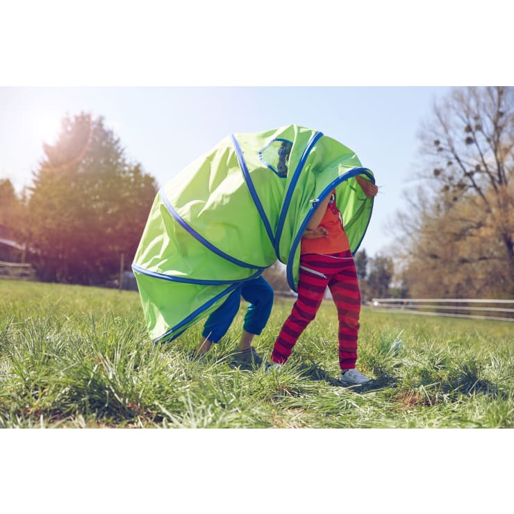 Zwei Kinder stehen auf einer Wiese im Freien, die teilweise von einem großen, grünen, zusammenklappbaren Spieltunnel mit blauer Umrandung bedeckt ist. Im Hintergrund sind Bäume und ein sonniger Himmel zu sehen.