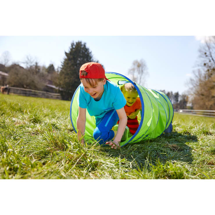 Zwei Kinder krabbeln durch einen grünen Spieltunnel auf einer Wiese. Das Kind vorne trägt eine rote Mütze und ein blaues Hemd, während das Kind hinten ein gelbes Hemd trägt. Im Hintergrund sind Bäume und ein Zaun zu sehen.