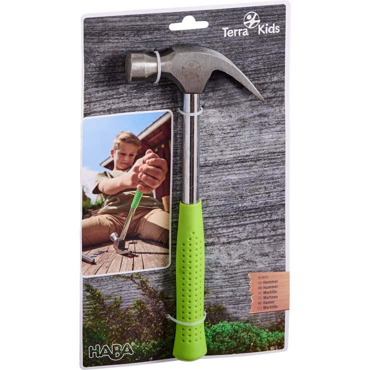 Ein verpackter Terra Kids Hammer mit grünem Stiel, der auf einer Papprückwand mit dem Foto eines Kindes abgebildet ist, das den Hammer benutzt, um im Freien einen Nagel in Holz zu schlagen. Das HABA-Logo ist am unteren Rand zu sehen.
