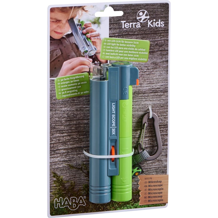 Ein verpacktes Terra Kids Mikroskop und ein Lichtmikroskop von HABA sind auf einer Kartonunterlage befestigt. Die Verpackung enthält einen Karabinerhaken, Produktdetails und ein Foto eines Kindes, das das Mikroskop im Freien benutzt.