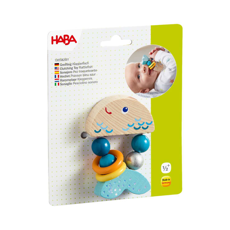 Ein Babyspielzeug der Marke HABA mit dem Namen Clutching Toy Rattlefish ist auf einer Karte verpackt. Das Spielzeug hat die Form eines Fisches mit Ringen aus Holz, Metall und Kunststoff. Auf der Verpackung ist ein Foto eines Babys abgebildet, das das Spielzeug hält.