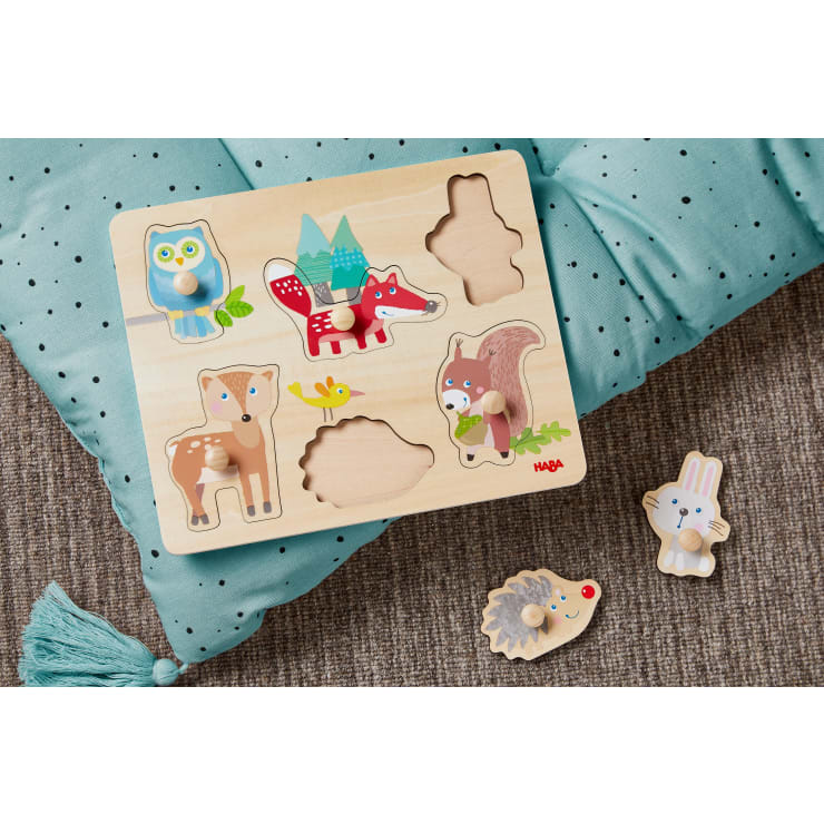 Ein hölzernes Tierpuzzle mit geformten Teilen einer Eule, eines Fuchses, eines Rehs, eines Eichhörnchens, eines Igels und eines Kaninchens liegt auf einem hellblauen Kissen auf einem Teppichboden. Zwei Teile sind herausgenommen und liegen neben dem Puzzlebrett.
