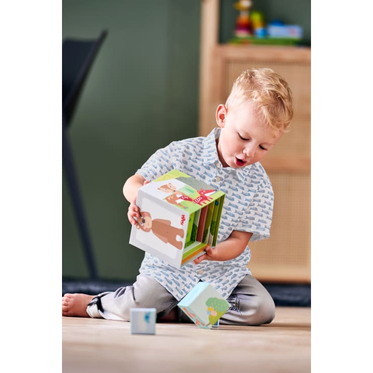 Ein kleines Kind mit blondem Haar sitzt auf dem Boden, hält bunte Stapelklötze mit Tiermotiven in der Hand und untersucht sie in einem Raum mit Holzfußboden und grüner Wand.