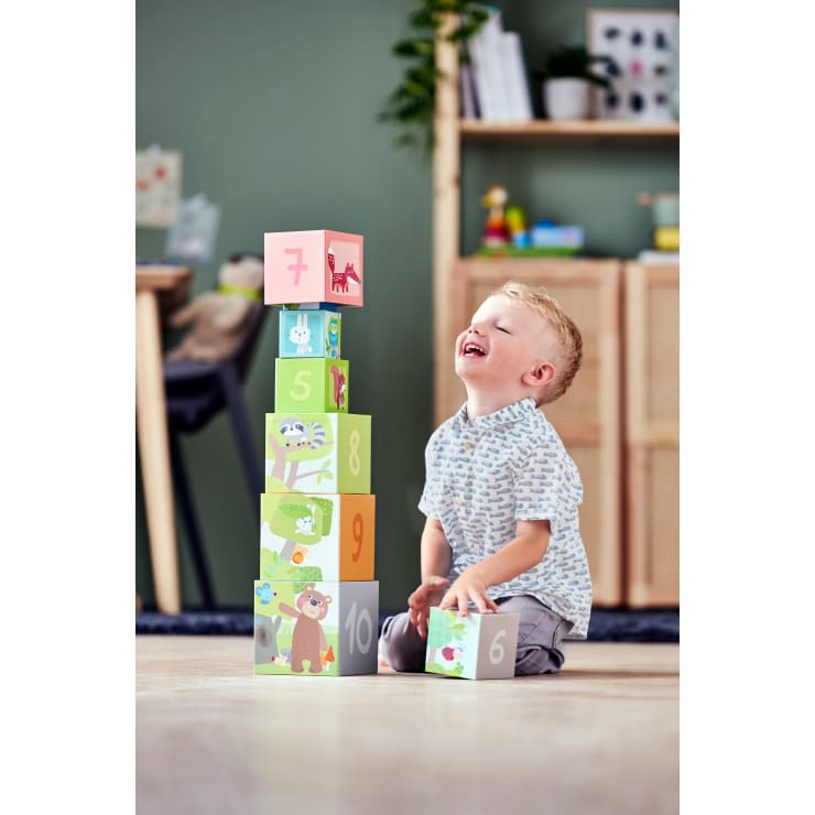 Ein kleines Kind kniet lächelnd auf dem Boden und stapelt bunte, nummerierte und illustrierte Blöcke zu einem Turm. Der Hintergrund zeigt ein aufgeräumtes Zimmer mit Regalen, Pflanzen und Spielzeug.