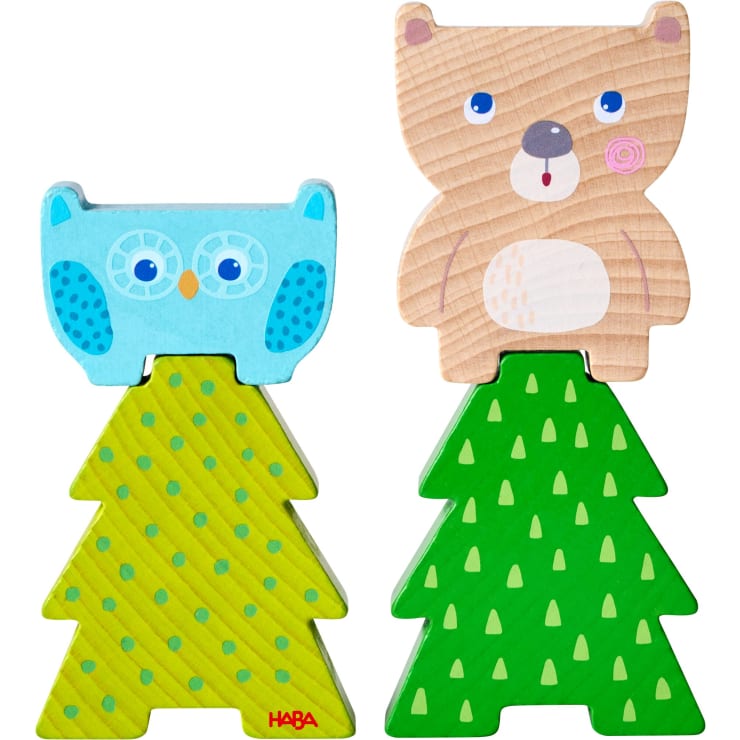 Zwei Holzspielzeuge sind abgebildet: eine blaue Eule, die auf einem gelb-grünen Baum sitzt, und ein brauner Bär mit weißem Bauch, der auf einem grünen Baum steht. Die Spielzeuge haben einfache Gesichtszüge und sind mit Mustern verziert.