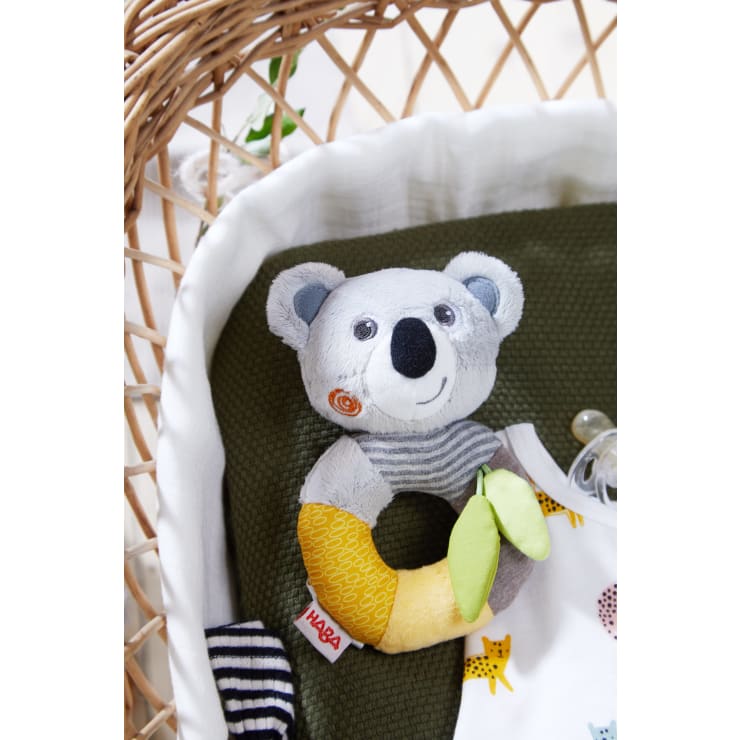 Eine plüschige Babyrassel in Form eines Koalas mit einem gestreiften Hals und einem gelben, grünen und beigen Ring liegt auf einer dunkelgrünen Decke in einem Weidenkorb. In der Nähe befinden sich ein Schnuller und Babykleidung mit Katzenmotiv.