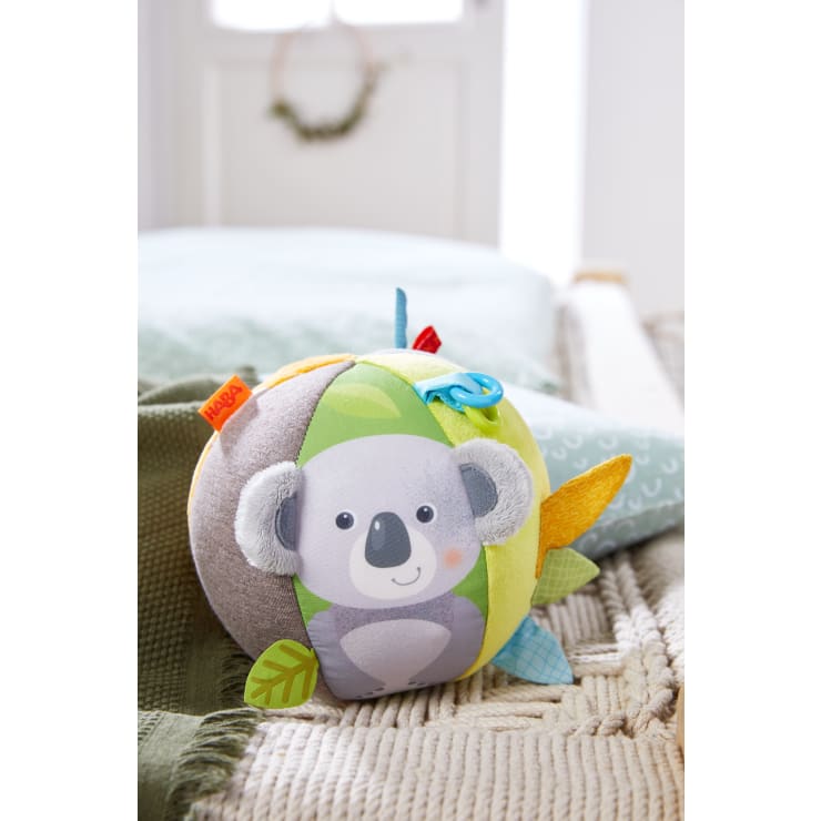 Ein weicher, farbenfroher Stoffball mit einer Koala-Illustration auf einer Seite, strukturierten Stoffanhängern und einer kleinen blauen Schleife, die auf einer grünen Strickdecke in einem gut beleuchteten Raum ruht.