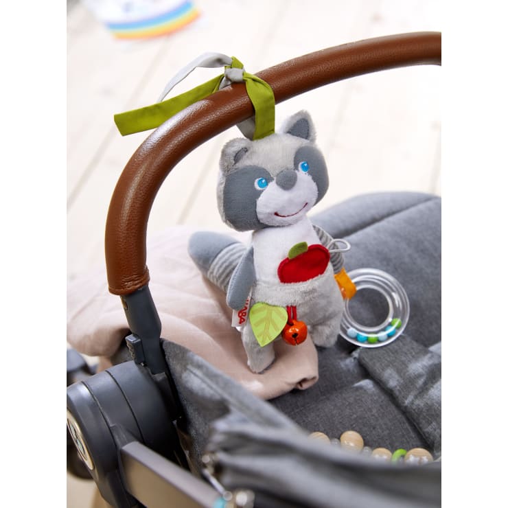 Ein grauer Plüschwaschbär mit einem roten Apfel und Blättern hängt an einem grünen Band an einem braunen Kinderwagengriff über einem grauen Sitz. Eine Plastikrassel und einige Holzperlen sind ebenfalls zu sehen.