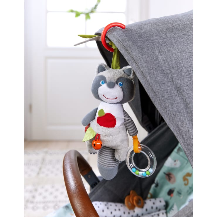 Ein grauer Plüschwaschbär mit einem roten Apfel auf dem Bauch hängt an einem roten Kunststoffring, der an einem grauen Kinderwagenverdeck befestigt ist. Das Spielzeug hat eine Glocke, ein Blatt und einen durchsichtigen Ring mit bunten Perlen.