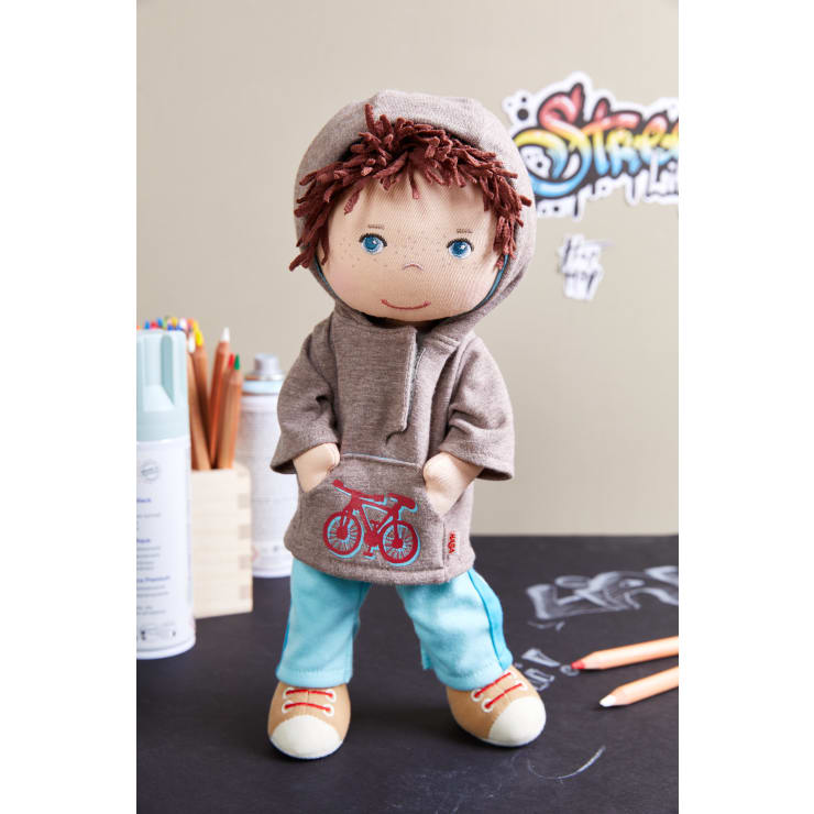 Eine Stoffpuppe mit braunen Haaren, blauen Augen und einem grauen Kapuzensweatshirt mit einer roten Fahrradgrafik steht auf einer schwarzen Fläche. Um sie herum liegen Buntstifte und Malutensilien, im Hintergrund ist Graffiti-Kunst zu sehen.