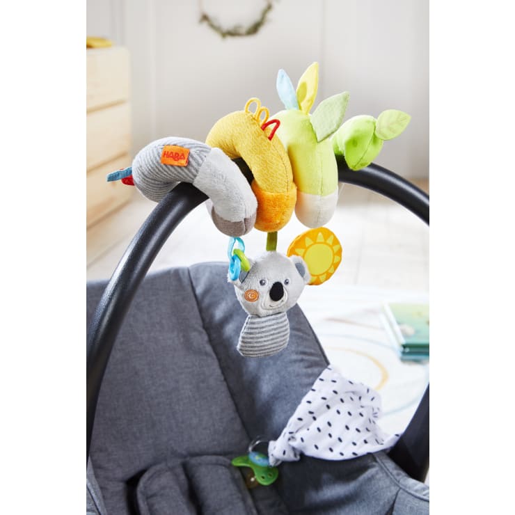Ein Griff für die Babyschale, an dem ein weiches, buntes Spiralspielzeug befestigt ist. Das Spielzeug zeigt Plüschtiere, darunter einen Koala, und hängende Elemente in Gelb, Grün und Grau. Ein Schnuller und ein gemustertes Tuch sind ebenfalls zu sehen.