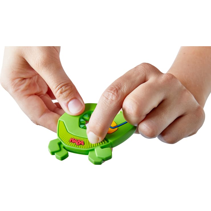 Eine Person benutzt ihre Hände, um ein kleines grünes Plastikspielzeug in Form eines Frosches mit dem Wort HABA darauf zu bedienen. Eine Hand hält das Spielzeug, während die andere einen zentralen Knopf drückt.